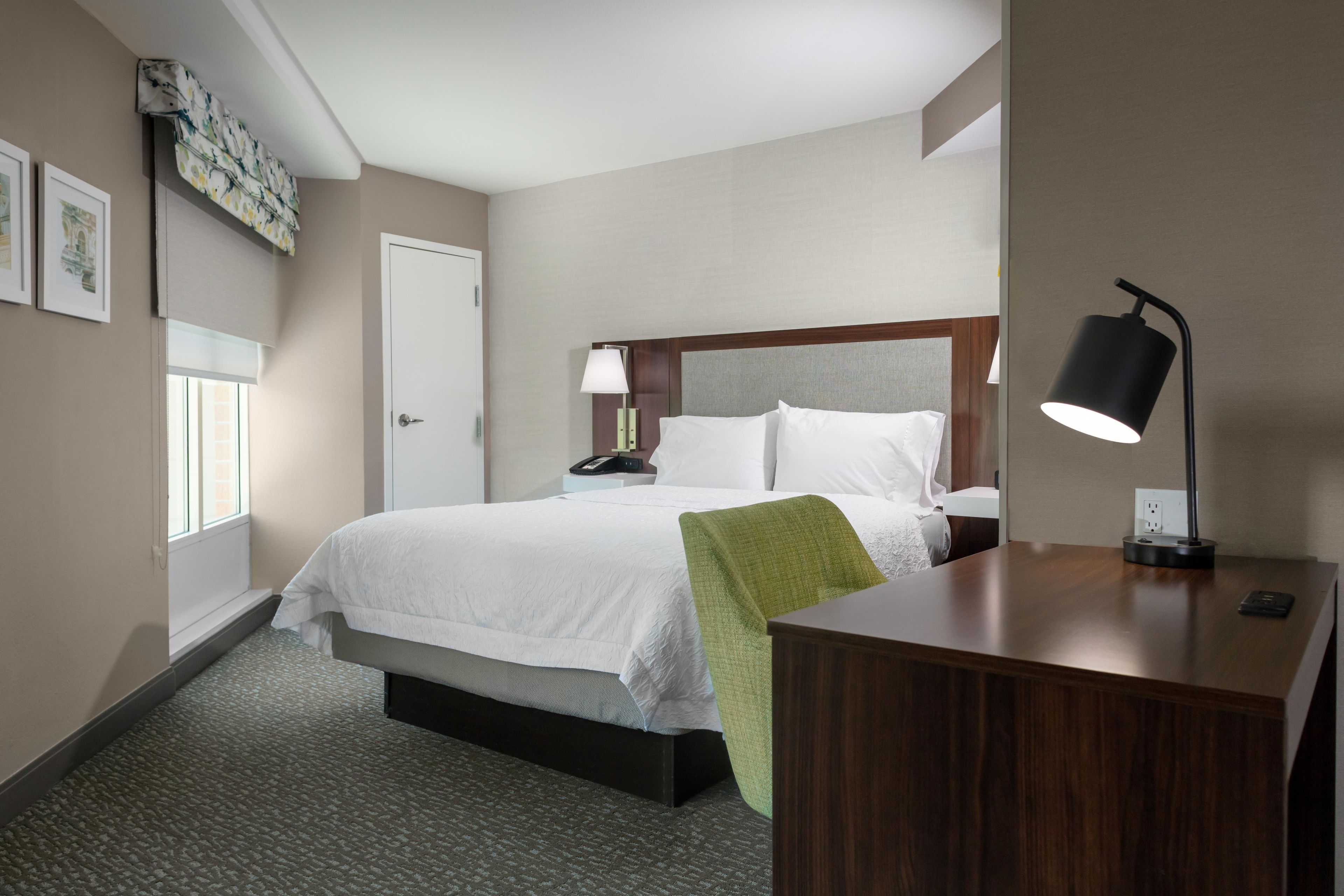 A hotel room with a bed, bedside table, lamp, and a window.