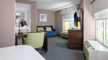 Una habitación de hotel con cama, escritorio con lámpara, sofá azul con cojines amarillos, una silla verde y una ventana con persianas.