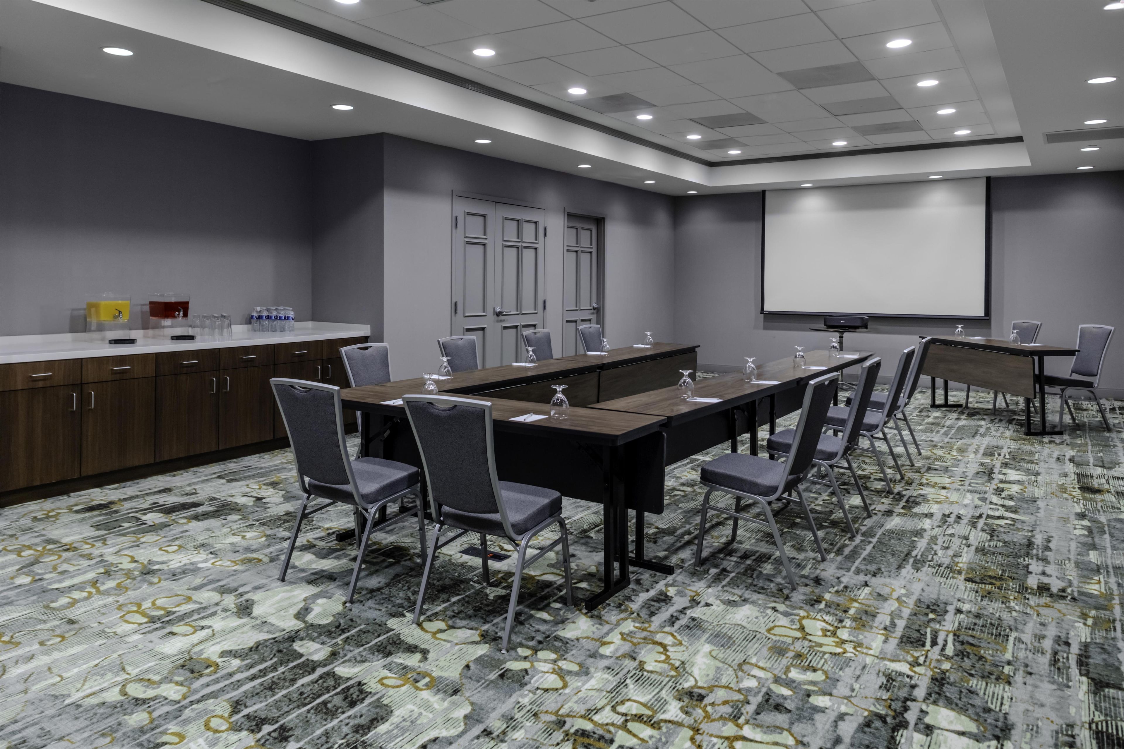 Sala de conferência com uma longa mesa, cadeiras, tela de projeção e um bebedouro.