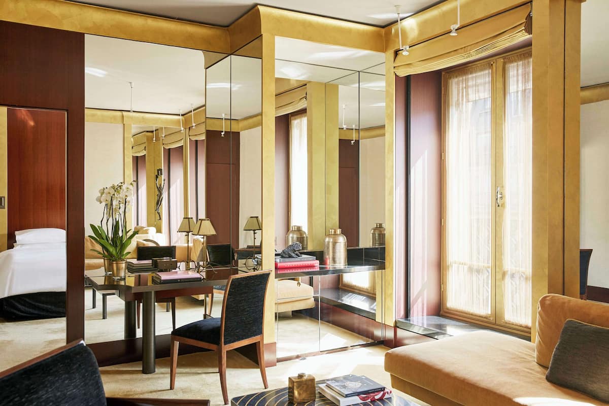 Ein Hotelzimmer mit einem großen Bett, einer Sitzecke inklusive Sofa und Sesseln, einem Esstisch mit Stühlen, einem großen Spiegel und einem Fenster mit Vorhängen.