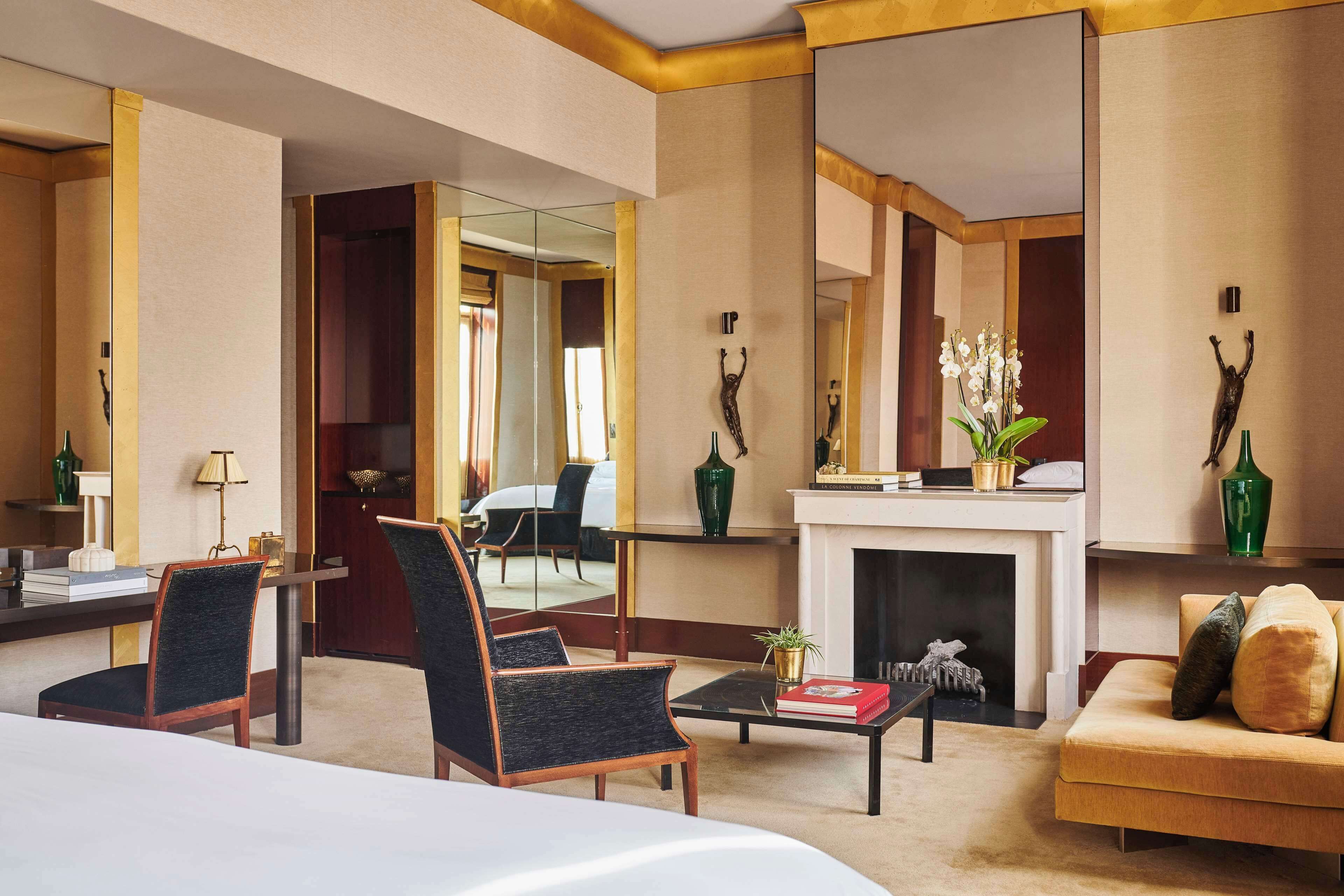 A hotel room with a large bed, a sofa, a coffee table, and a fireplace. There is a mirror on the wall and a plant on the table.