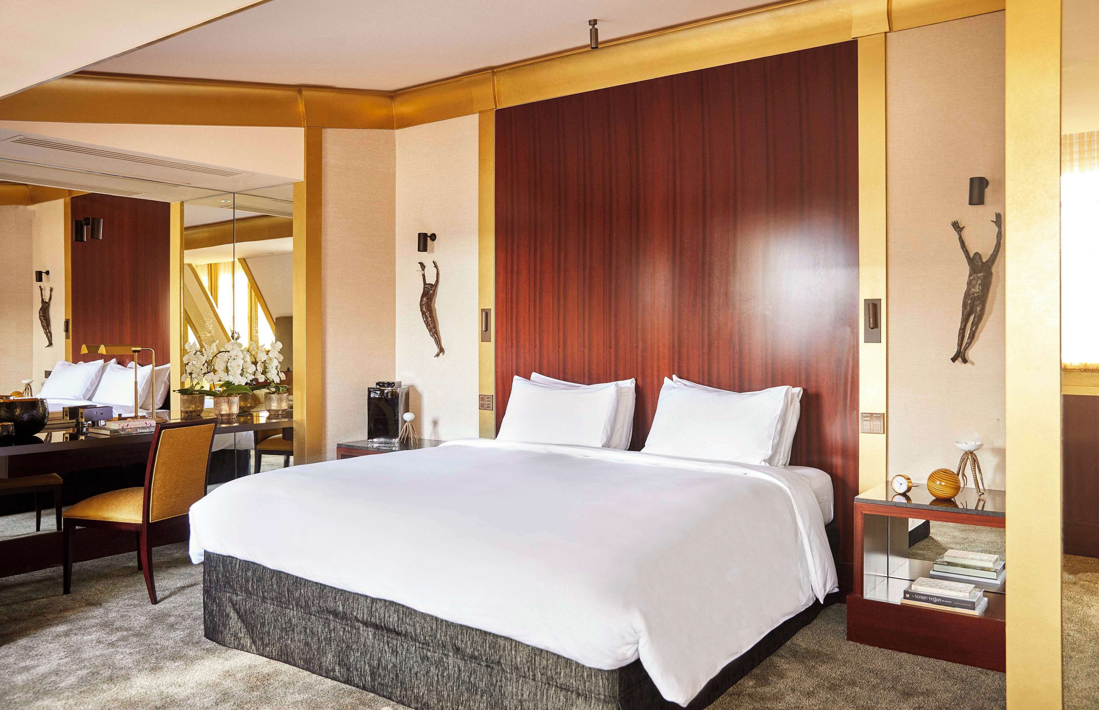 A hotel room with a large bed, a desk with a chair, a bedside table with books, and a mirror.