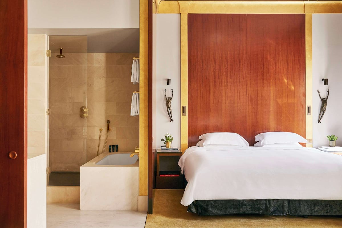 Ein modernes Hotelzimmer mit einem großen Bett, einem Holz-Kopfteil, einer Duschkabine und einem kleinen Nachttisch mit Pflanze.