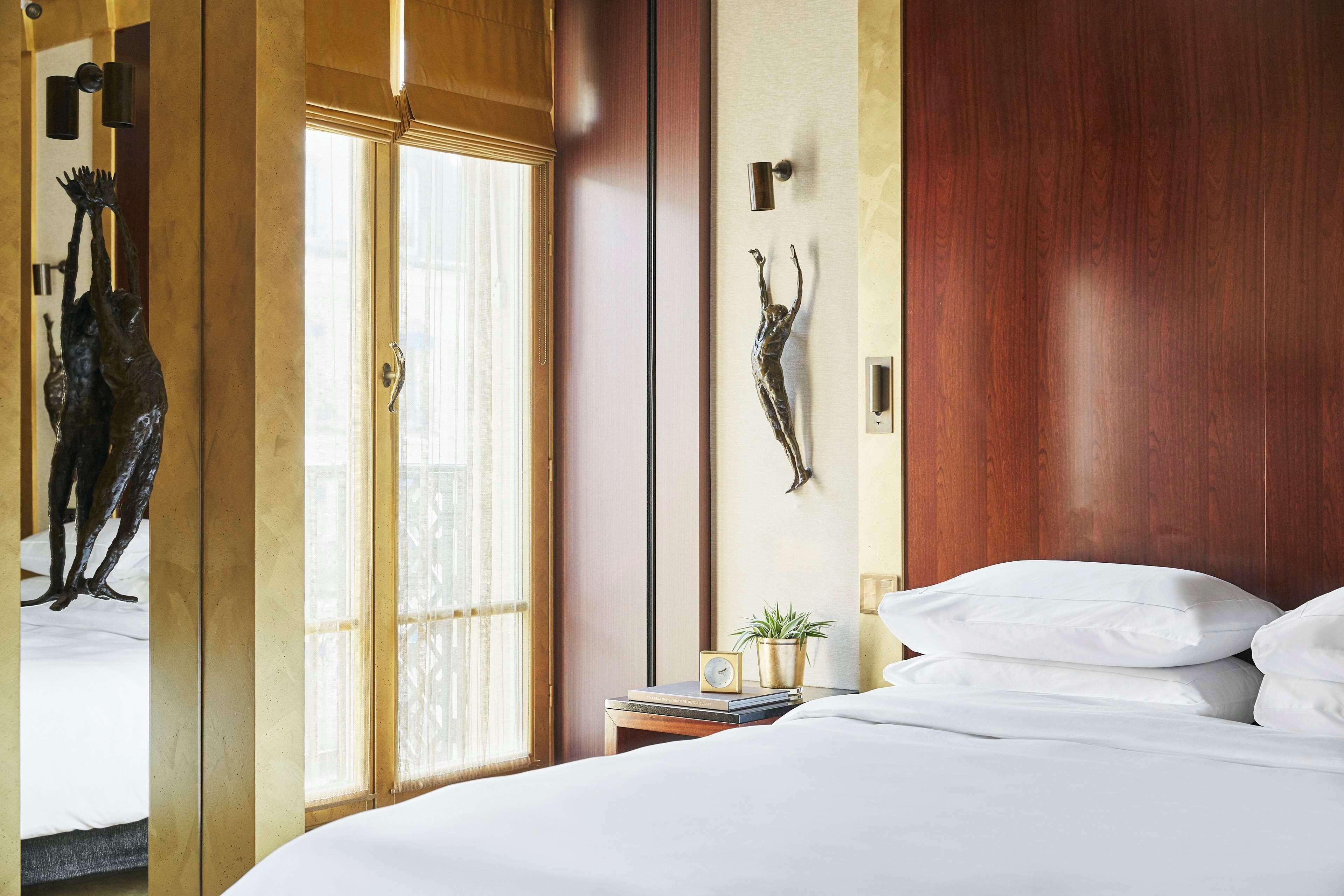 A hotel room with a bed, a nightstand with a clock and a plant, a wooden door, and a wall-mounted sculpture.