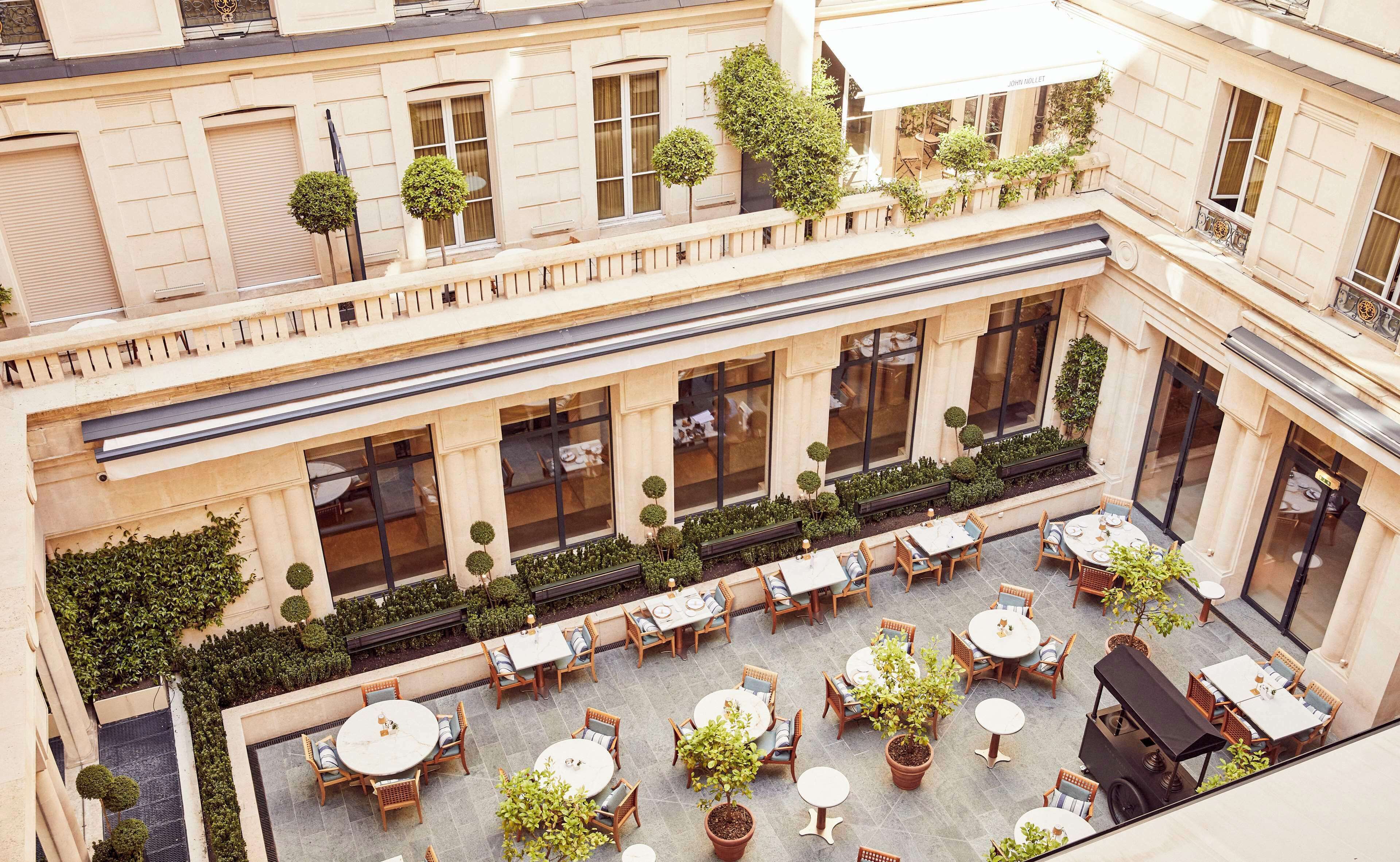Un patio con varias mesas y sillas, rodeado de follaje y un edificio con ventanas grandes.