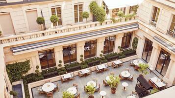 Un patio con varias mesas y sillas, rodeado de follaje y un edificio con ventanas grandes.