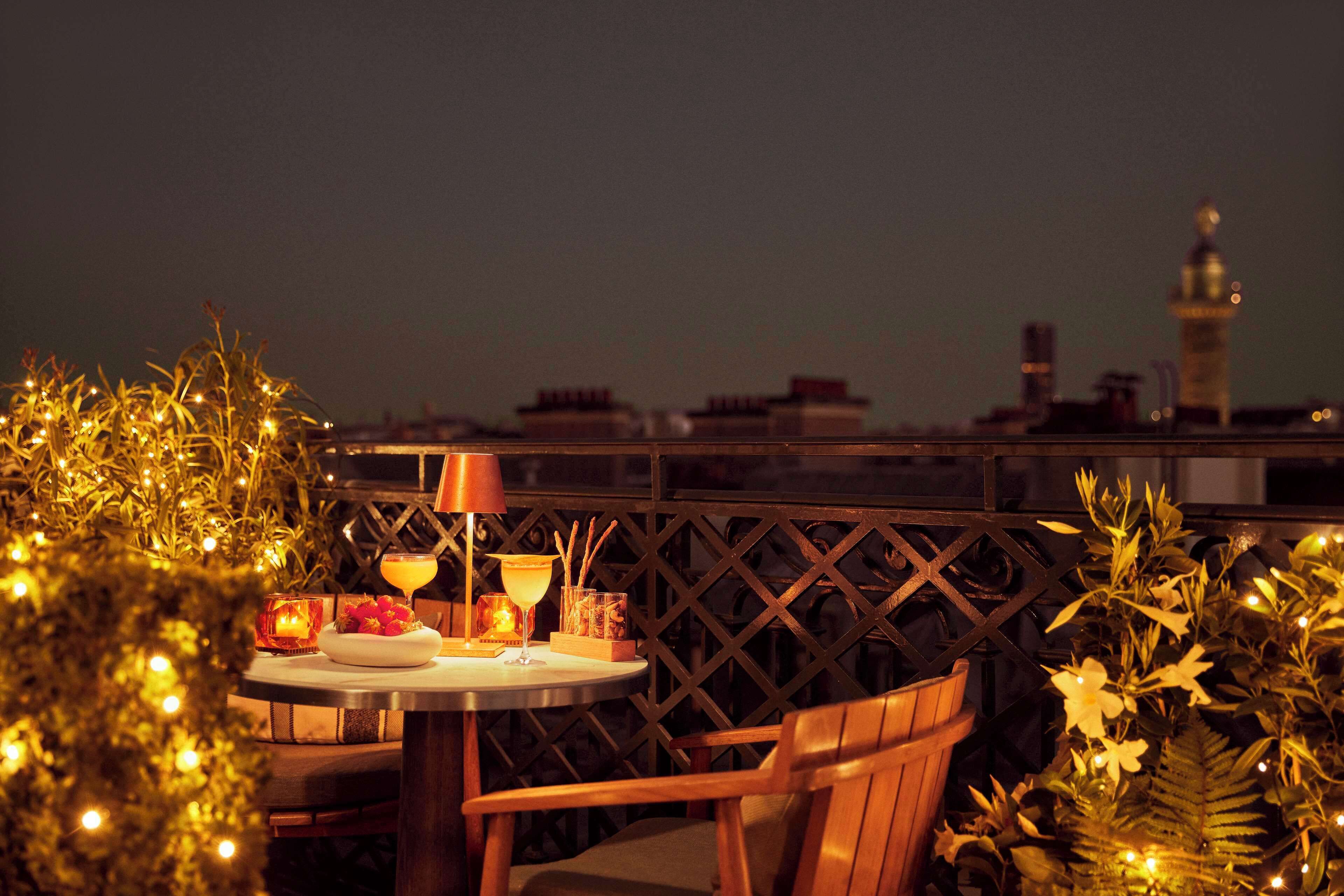 Una mesa dispuesta con una bebida, una vela y un bol de fruta, rodeada de plantas y una valla decorativa.