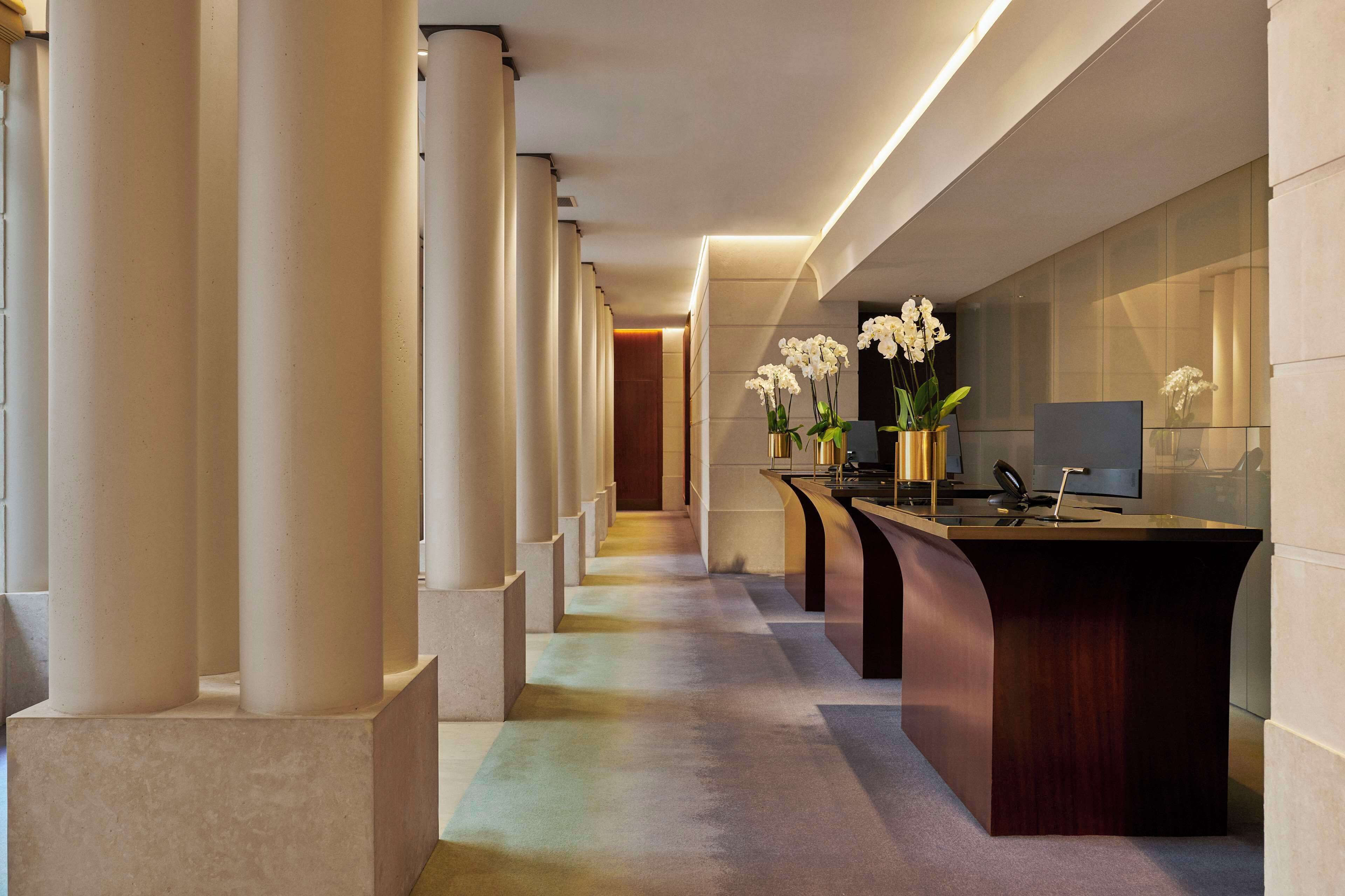 A modern hotel lobby with a long corridor, large columns, a reception desk with a computer, and white orchids.
