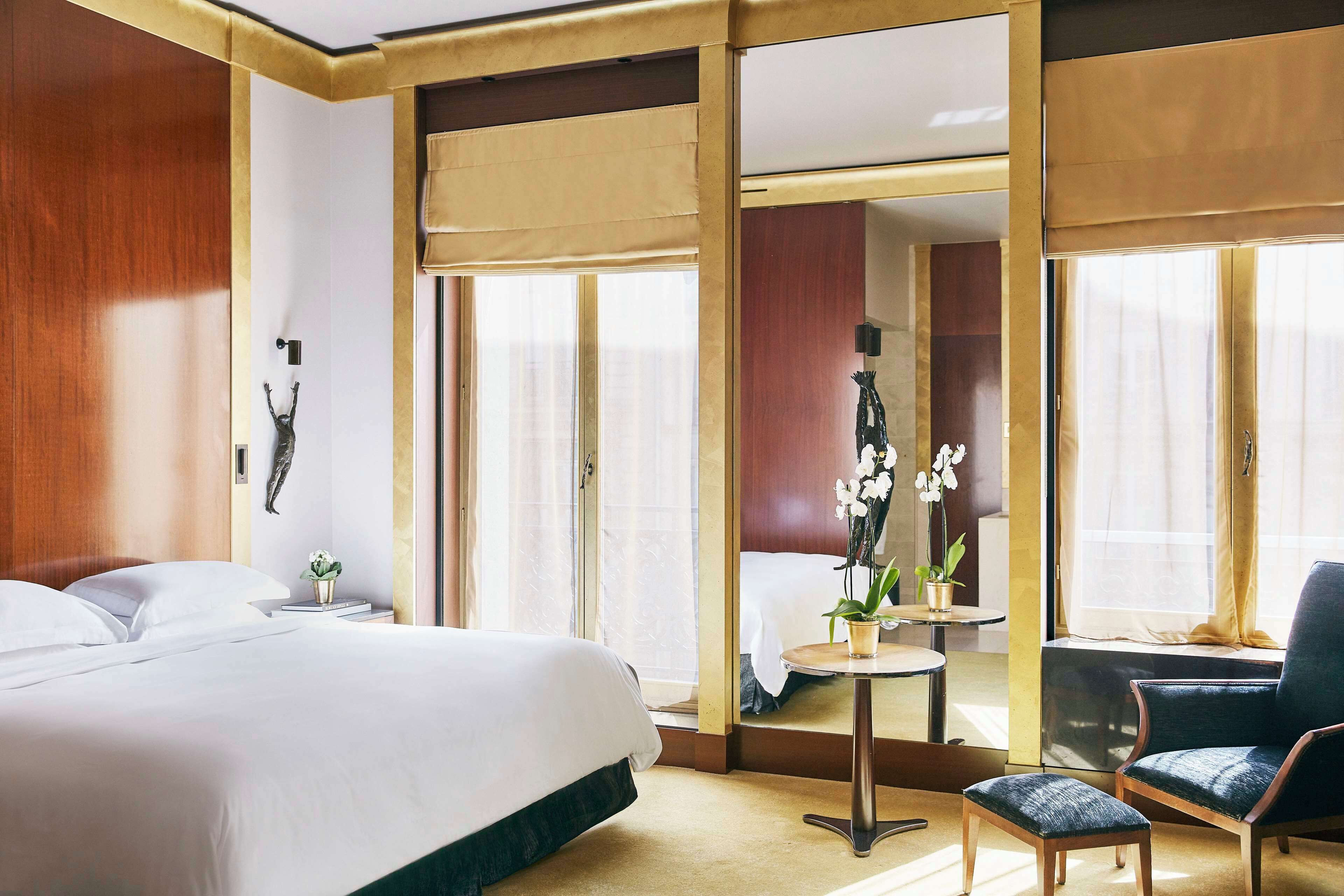 Habitación de hotel con una cama grande, una mesita con una planta, una silla y vistas a otra habitación a través de una puerta corredera.