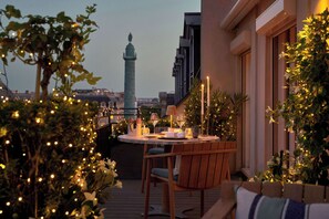 Un área de comedor al aire libre con una mesa redonda, sillas y luces colgantes. Se ve una torre alta al fondo.