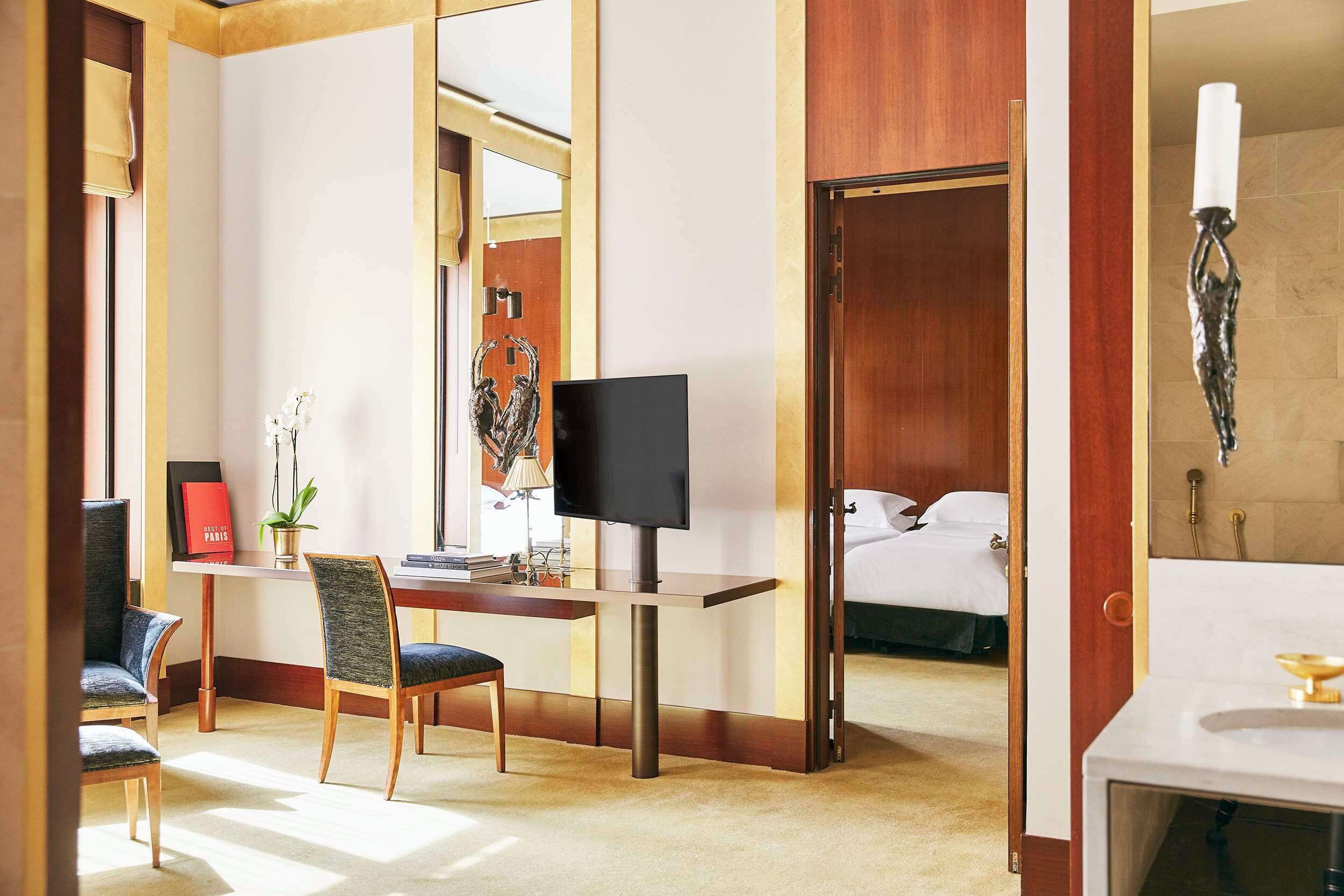 A hotel room with a desk, chair, television, bed, and a bathroom with a mirror and sink.