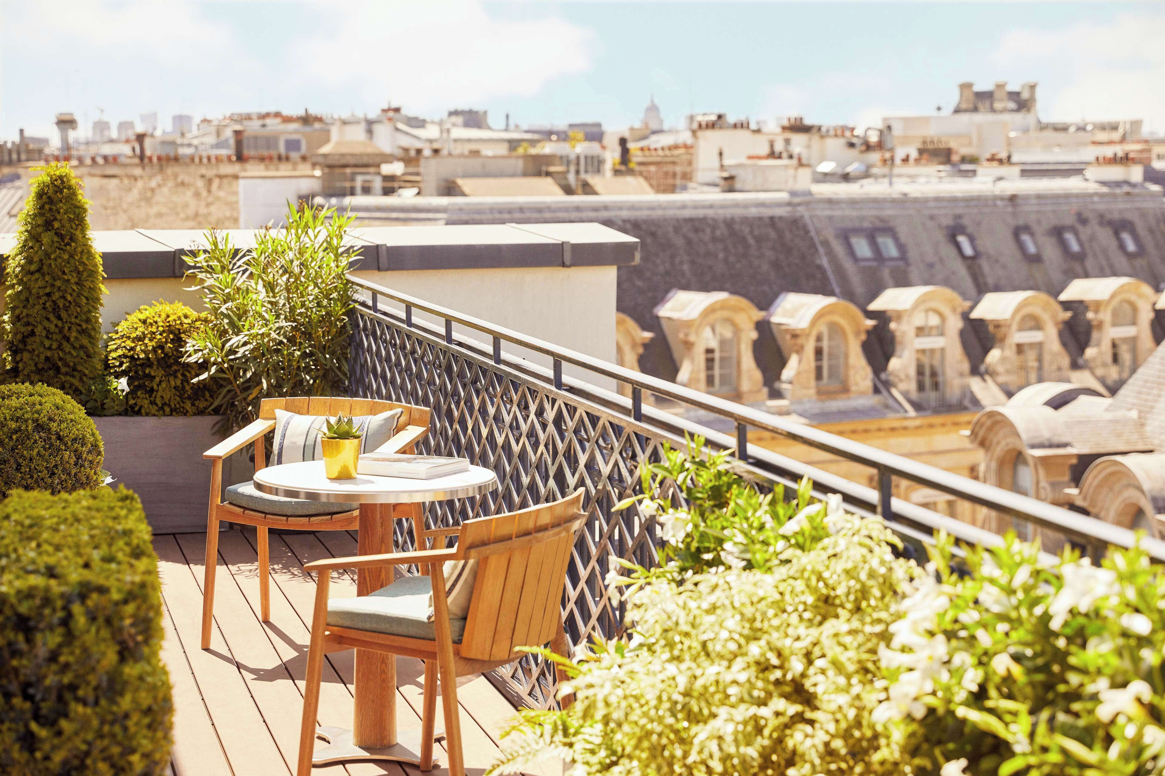 A rooftop terrace with a wooden table and chairs, surrounded by greenery and overlooking a cityscape with historical architecture.