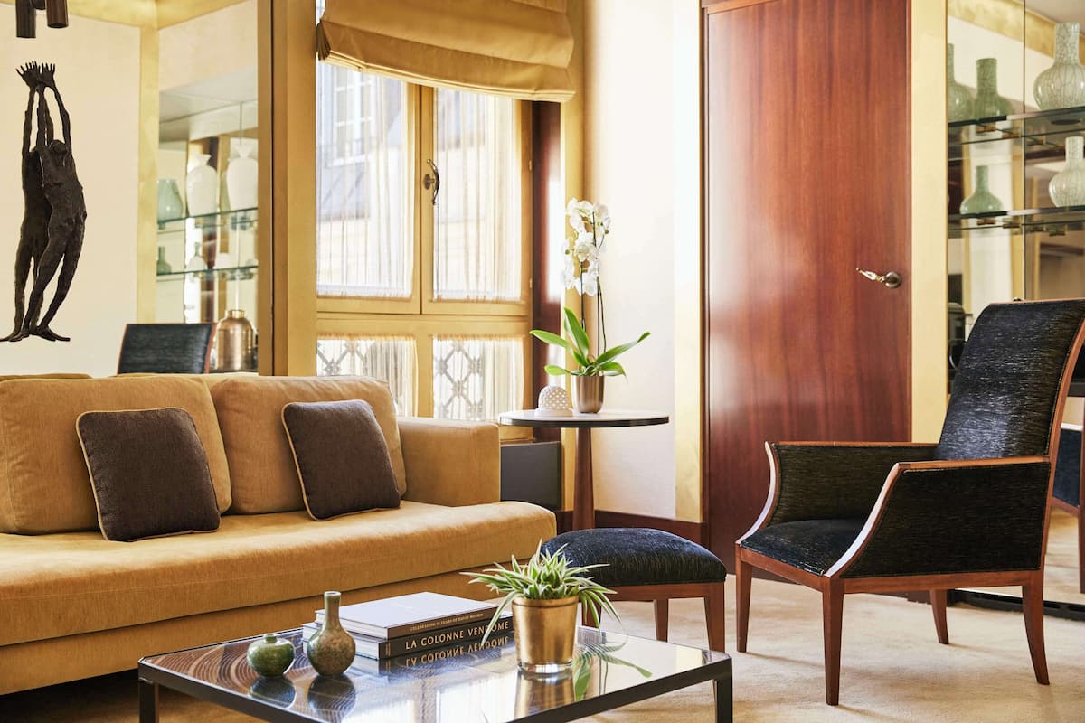 Ein Wohnzimmer mit Sofa, Sessel und Couchtisch. Es gibt ein Fenster mit Vorhängen, eine Tür und Regale mit Dekoration.