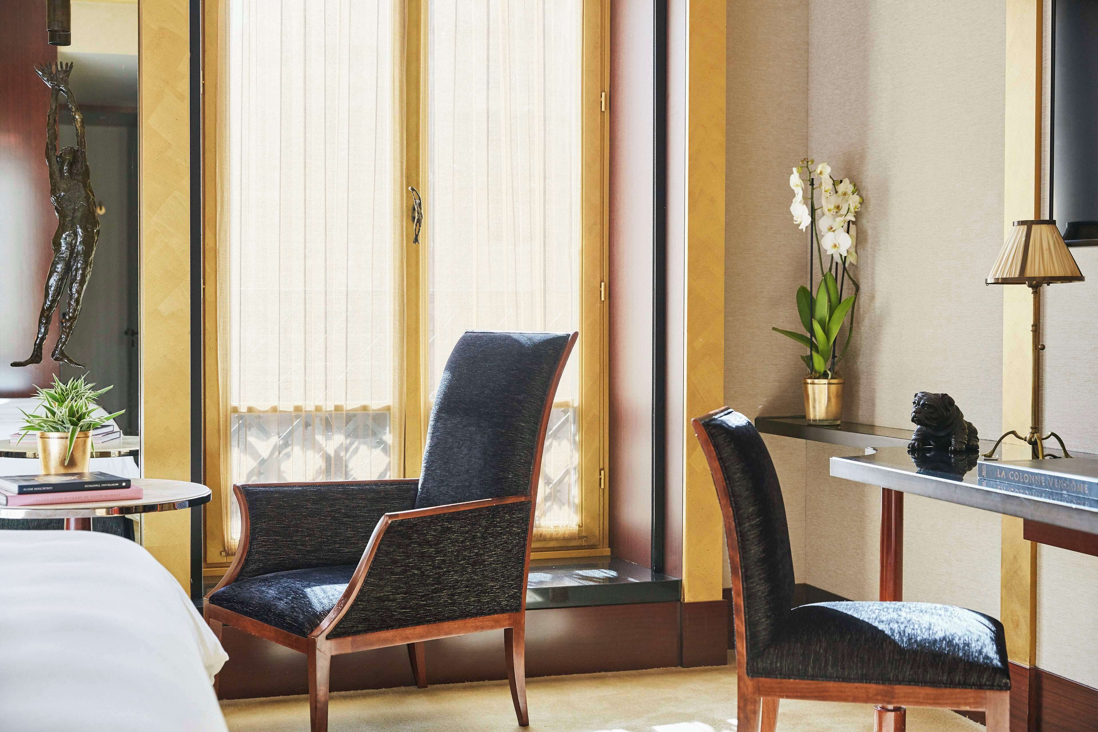 A hotel room with a large window, two chairs, a small table with a lamp, a potted plant, and a decorative sculpture.