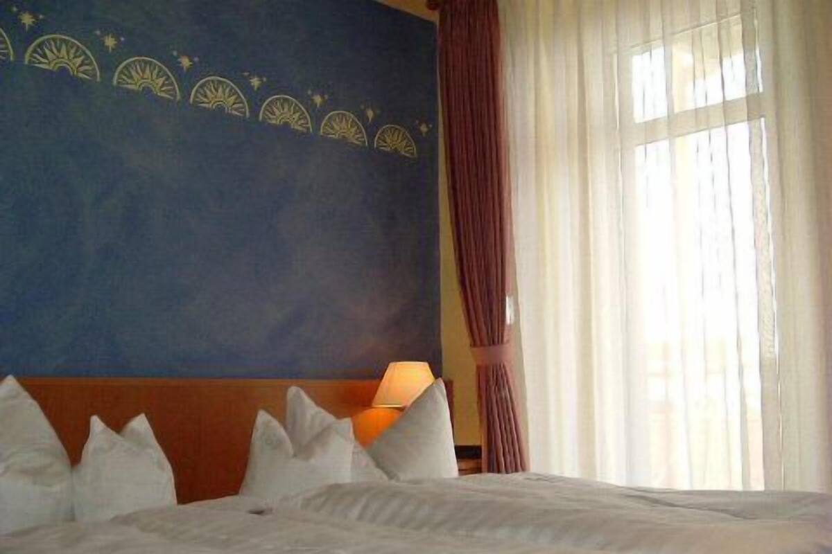 A hotel room with a large bed, a headboard with a decorative panel, a nightstand with a lamp, and a window with curtains.