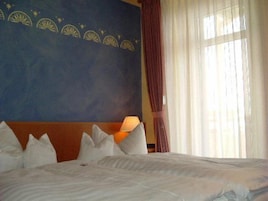 A hotel room with a large bed, a headboard with a decorative panel, a nightstand with a lamp, and a window with curtains.