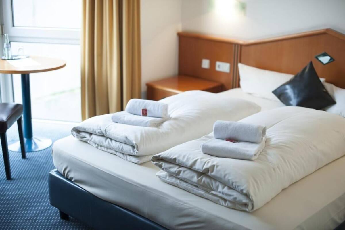 A hotel room with two beds, each with neatly folded towels, a wooden headboard, a small bedside table, and a window with curtains.