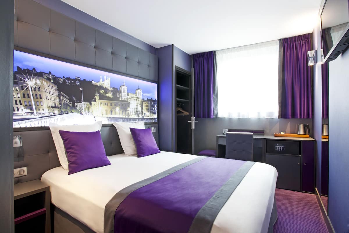 Une chambre d’hôtel moderne dotée d’un grand lit, offrant une vue sur le paysage urbain, d’un bureau avec une bouilloire et d’une armoire.