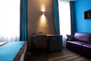 A hotel room with a bed, a desk with a chair, a small safe, a purple sofa, and a wall-mounted TV.
