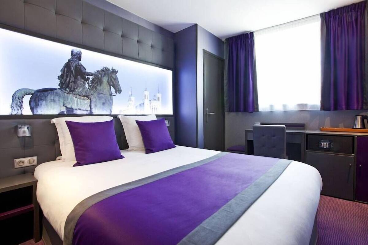 Une chambre d’hôtel avec un grand lit, une tête de lit ornée d’une œuvre d’art représentant un paysage urbain, un bureau avec une chaise et une fenêtre avec des rideaux violets.