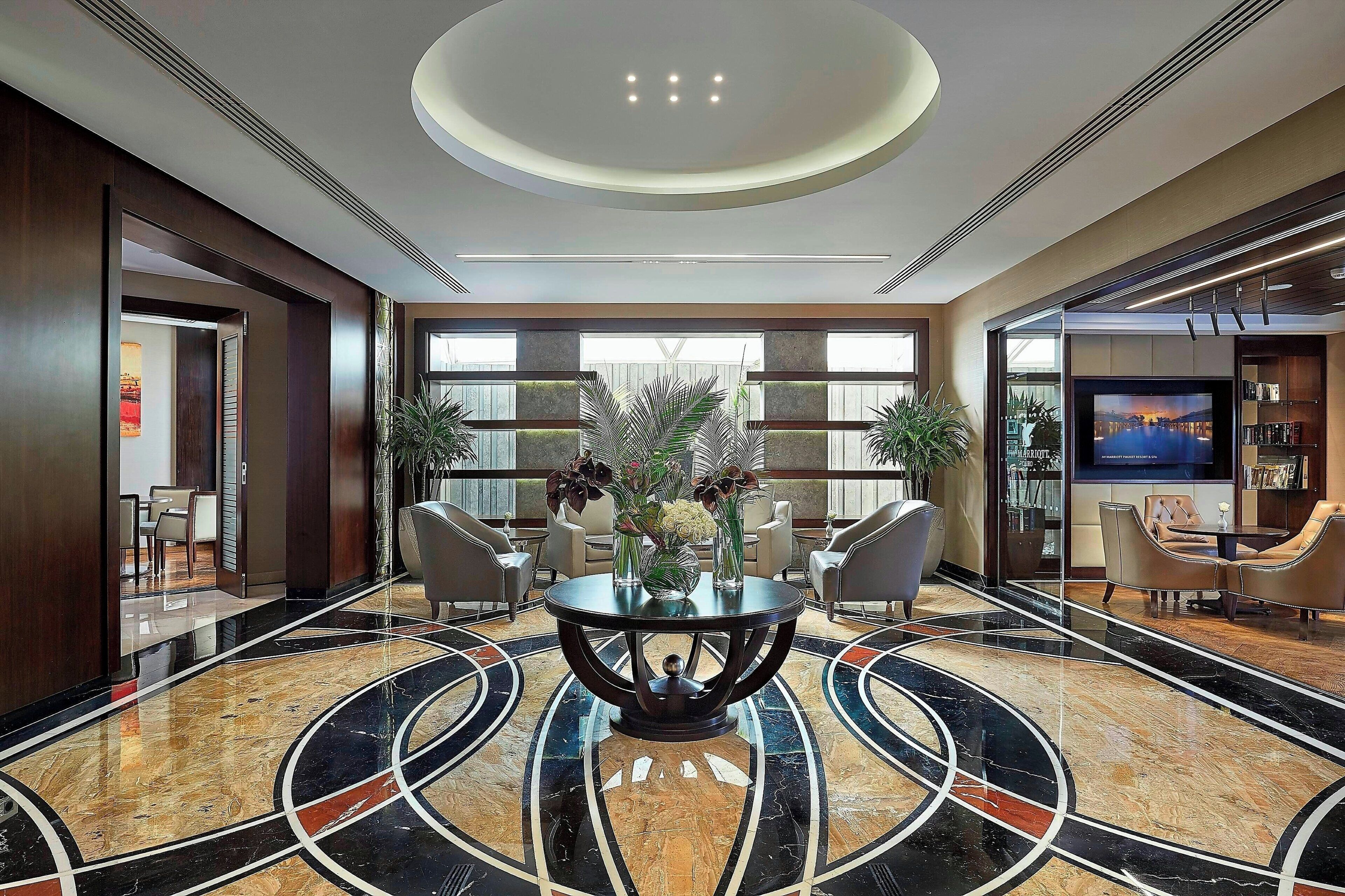 A modern lobby with a marble floor, a central round table with chairs, and a large TV screen.