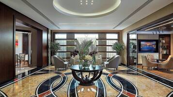 A modern lobby with a marble floor, a central round table with chairs, and a large TV screen.
