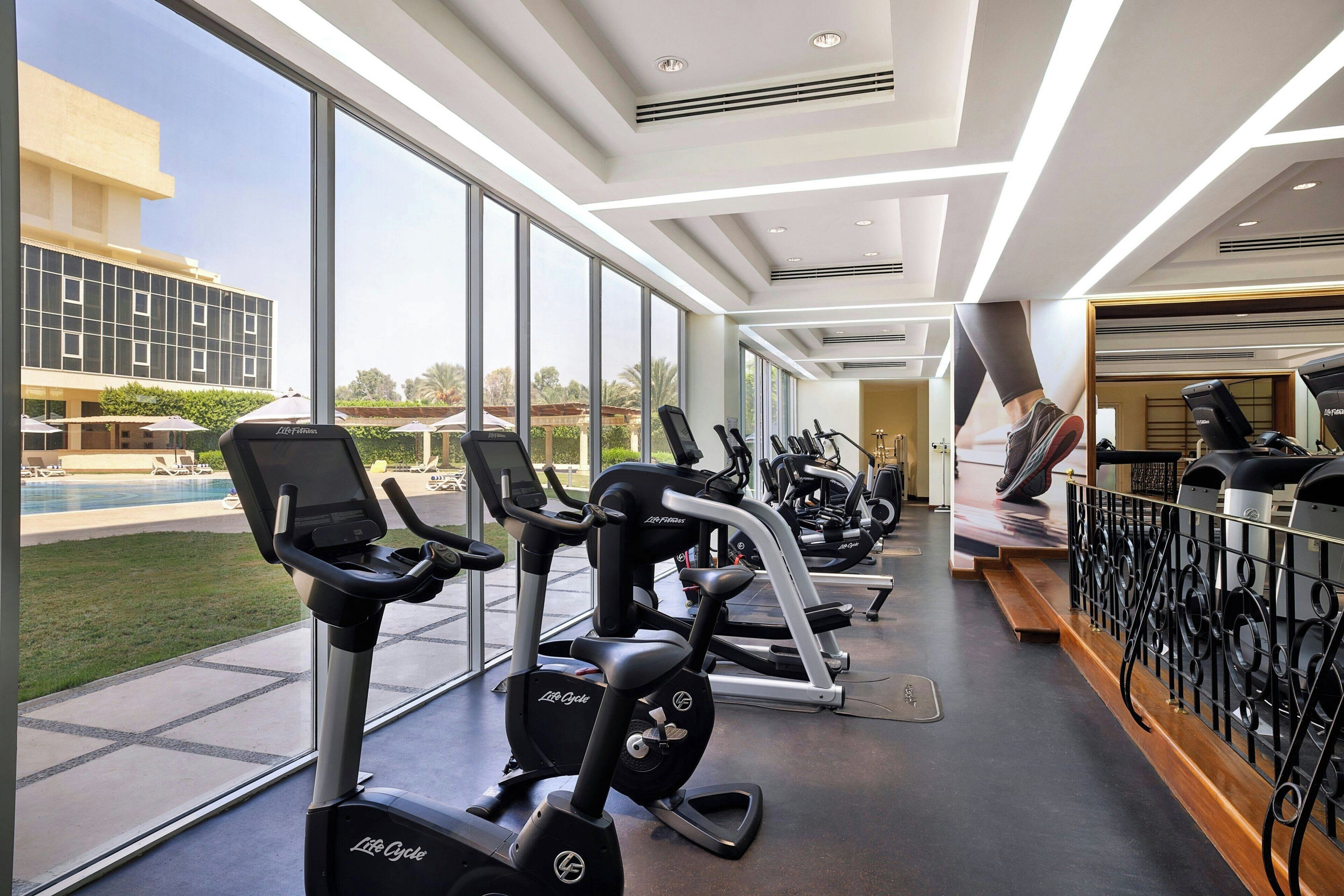 A gym with treadmills, ellipticals, and a pool in the background.