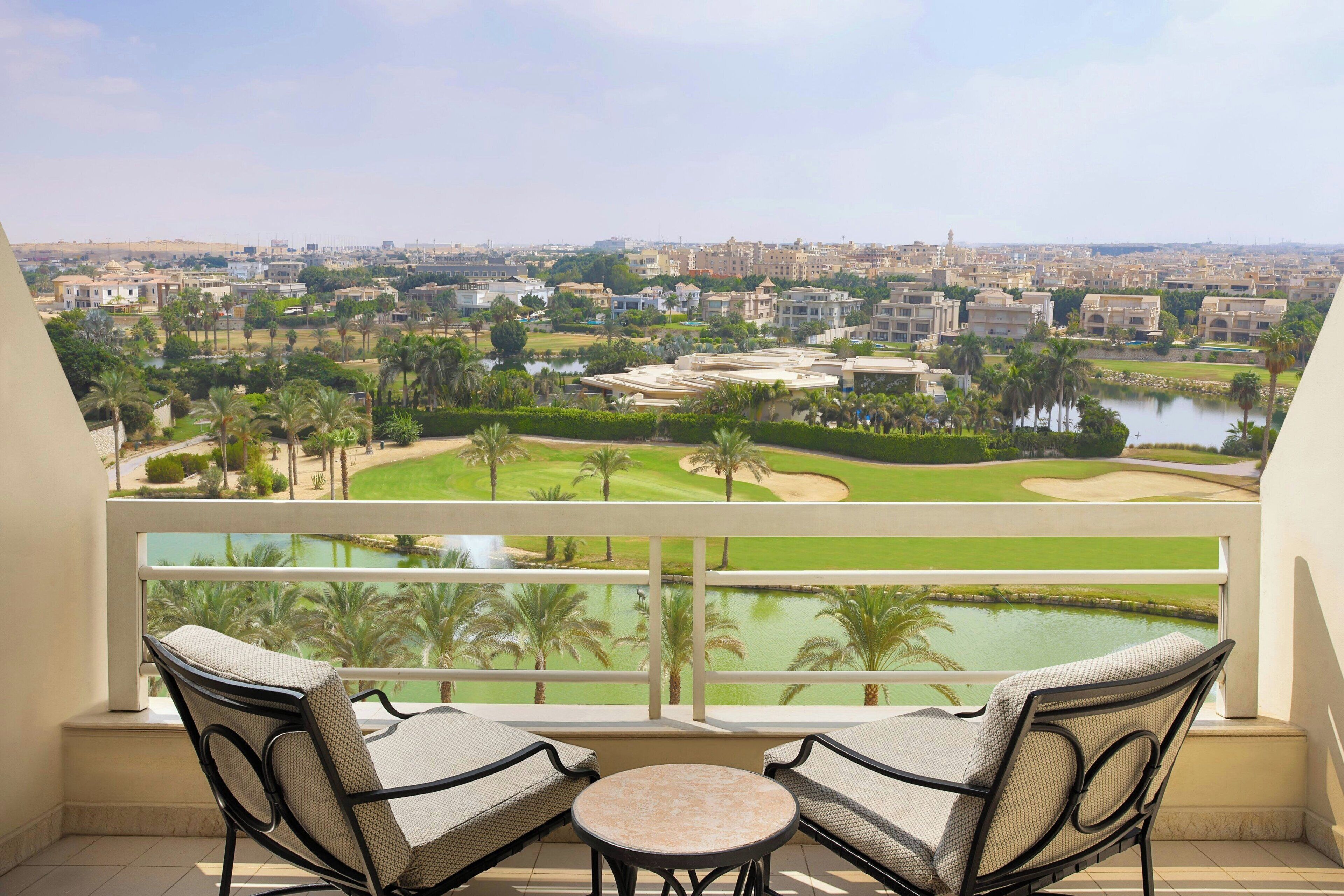 Un balcón con dos sillas de mimbre y una pequeña mesa, con vistas a un campo de golf y una vista urbana.