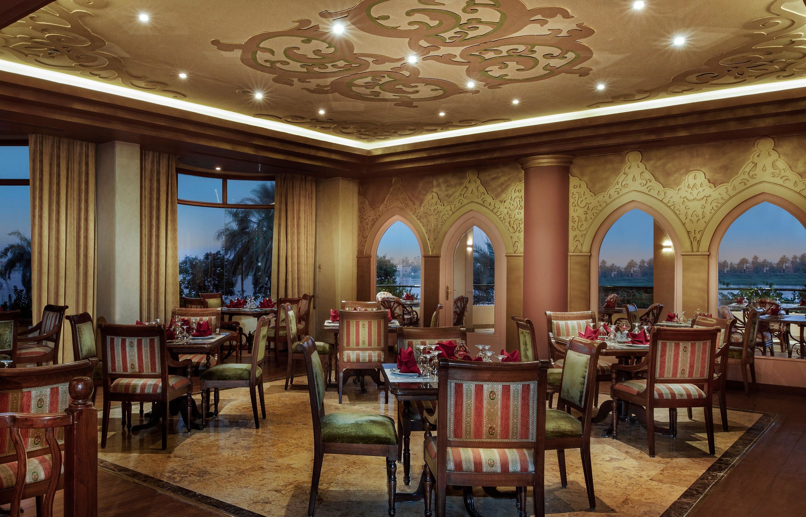 A dining area with ornate ceiling and arched windows, wooden tables and chairs, and a view of the outdoors.