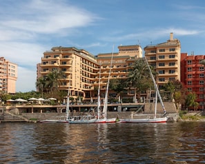 A hotel with multiple balconies, a waterfront view, and several boats docked nearby. - Steigenberger Nile Palace (Luxor)