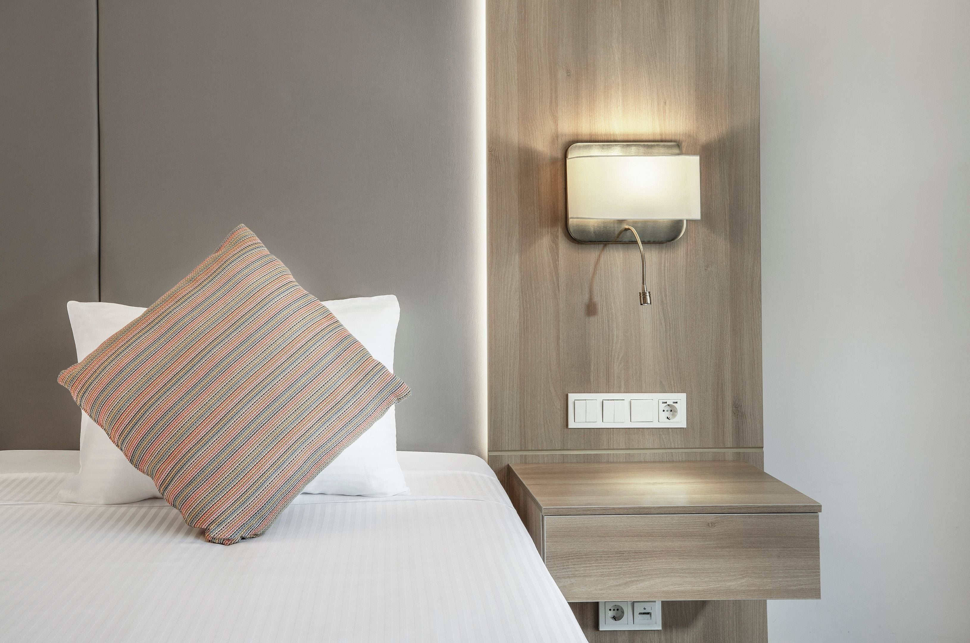 Una cama bien hecha con un almohadón a rayas, una mesita de noche con interruptor de luz y una lámpara fijada a la pared.