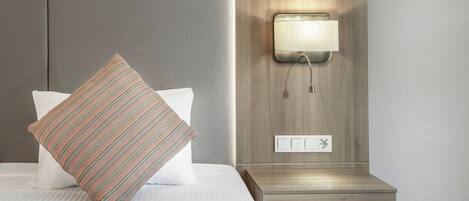 Una cama bien hecha con un almohadón a rayas, una mesita de noche con interruptor de luz y una lámpara fijada a la pared.