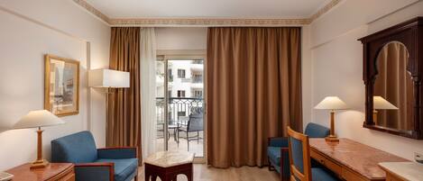 A hotel room with a bed, two blue armchairs, a wooden desk with a lamp, a small table, and a large mirror.