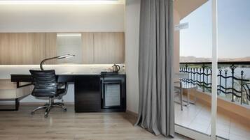 Una habitación de hotel moderna con un escritorio de madera, una silla de oficina negra, un refrigerador pequeño y vista a un cuerpo de agua a través de un balcón con barandilla de metal.
