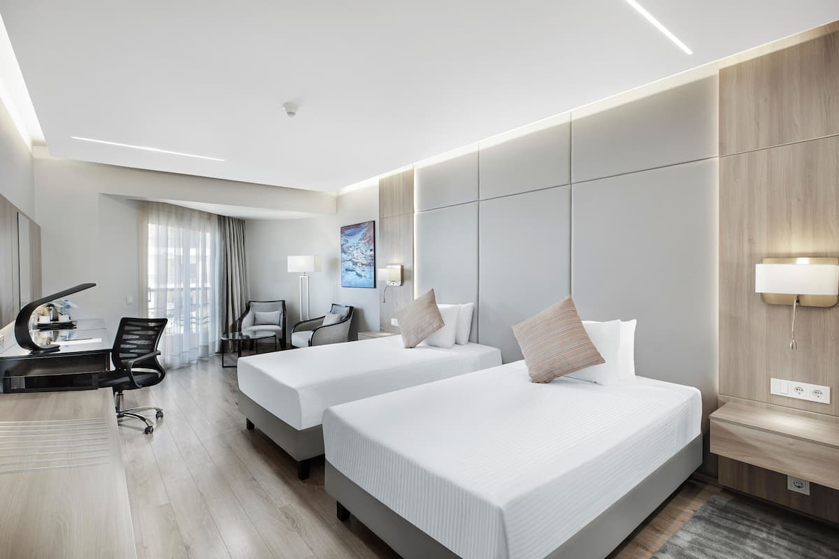 A modern hotel room with two beds, a desk with a chair, a TV, and a large window with curtains.