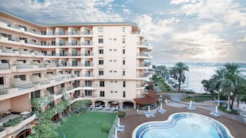 A multi-story hotel with a swimming pool, outdoor seating, and a view of the sea.