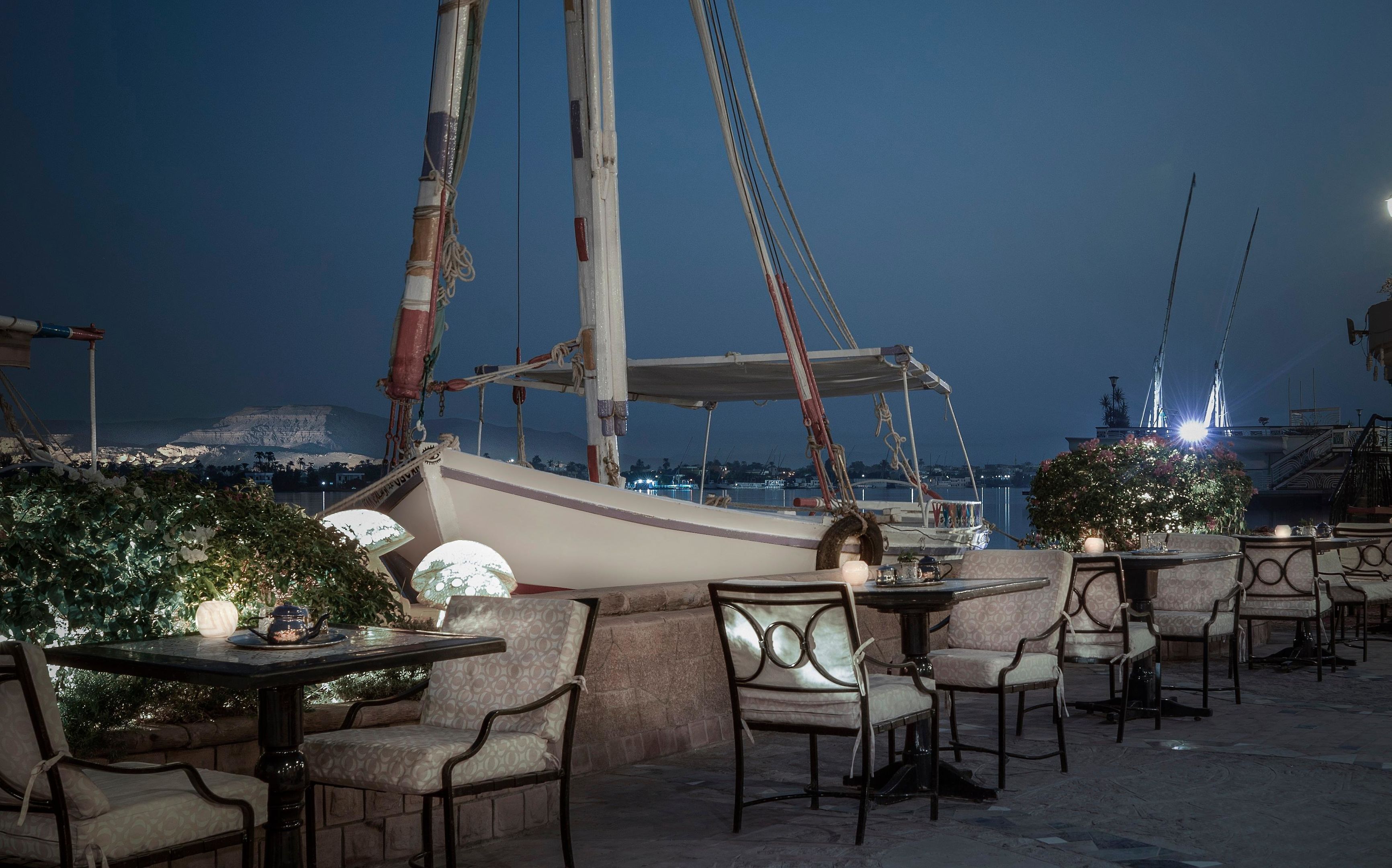 Un área de comedor al aire libre con mesas y sillas, un velero al fondo y un paisaje urbano al atardecer.