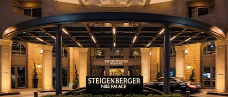 The entrance of Steigenberger Nile Palace with a curved canopy, illuminated signs, and a landscaped area.
