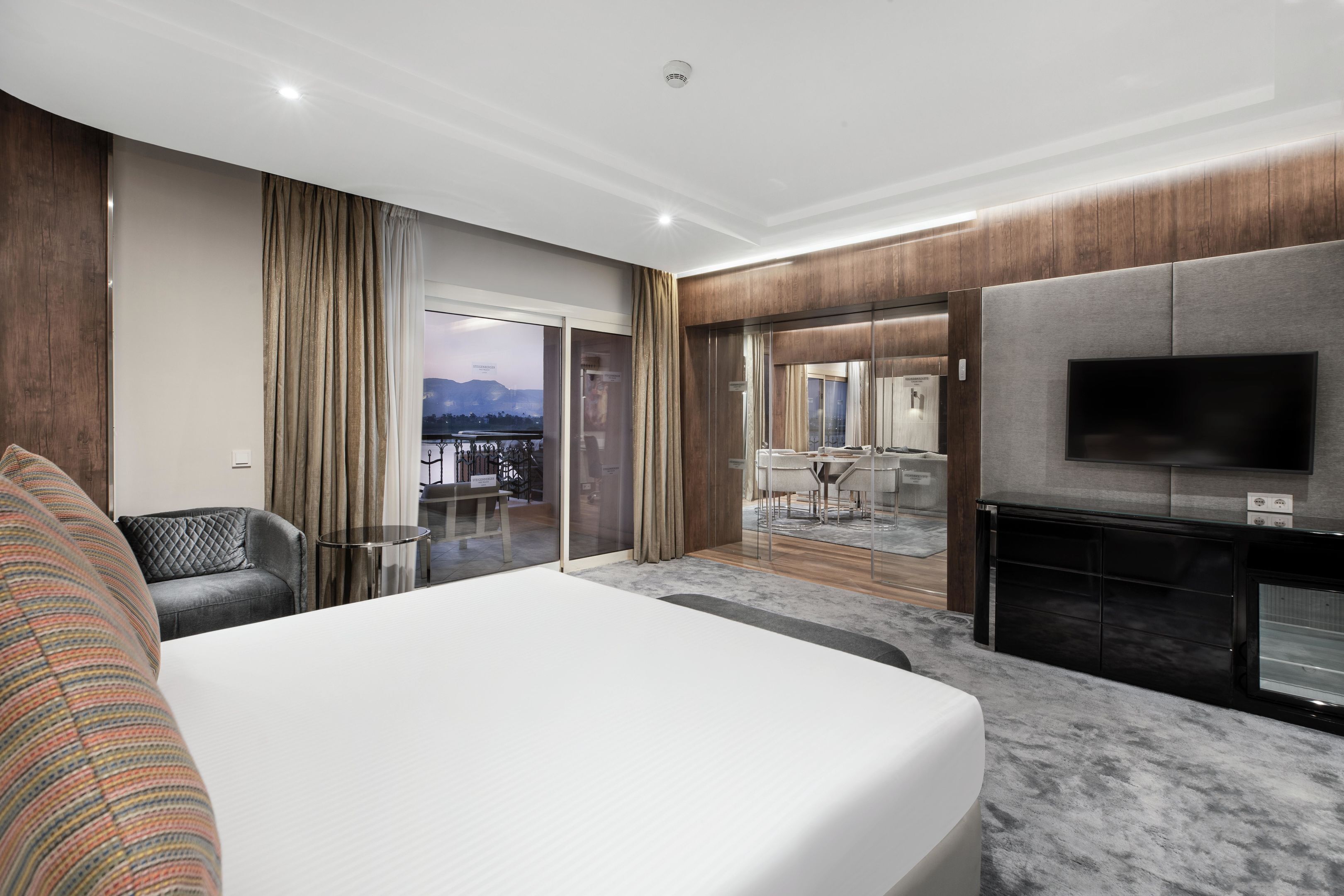 Una moderna camera d'hotel con un grande letto, una televisione a schermo piatto, un'area salotto con un tavolo e sedie, e vista sulla città dalla finestra.