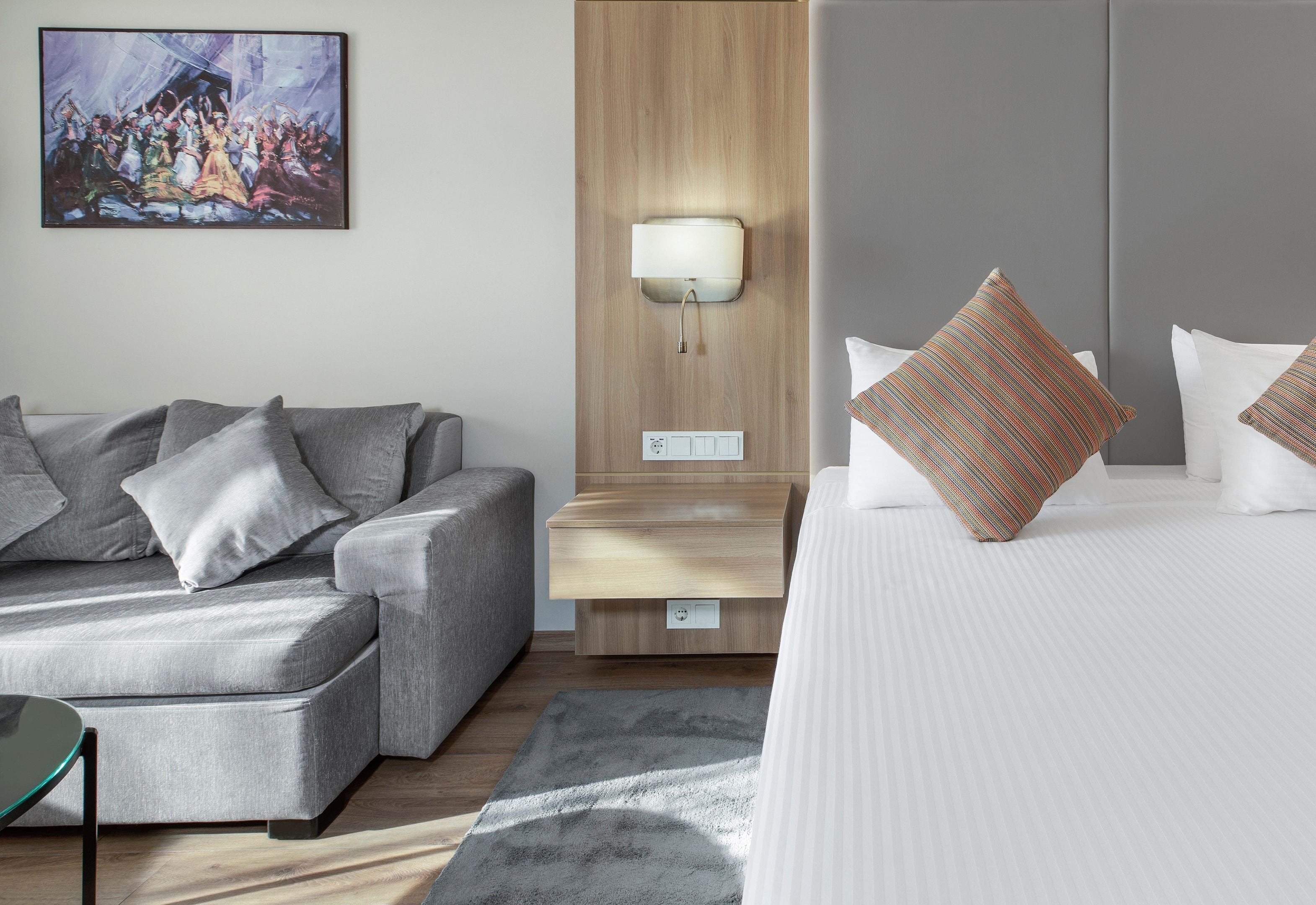 A modern hotel room with a grey sofa, a bed with striped pillows, a wooden nightstand with a lamp, and a painting on the wall.
