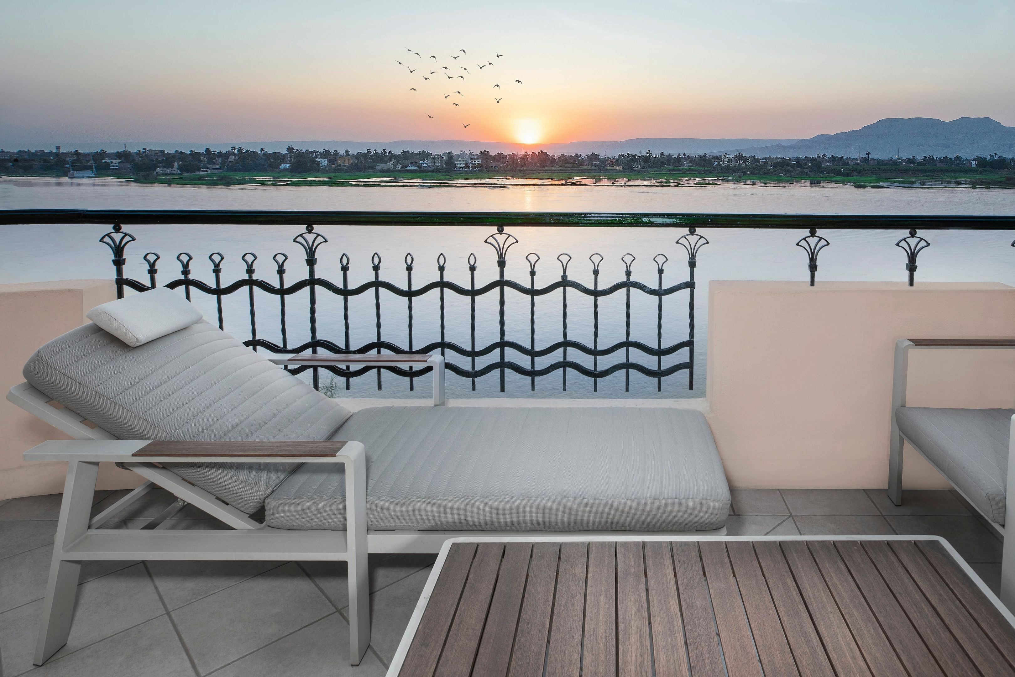 A balcony with a lounge chair and a small table, overlooking a body of water and a sunset.