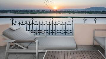 A balcony with a lounge chair and a small table, overlooking a body of water and a sunset.