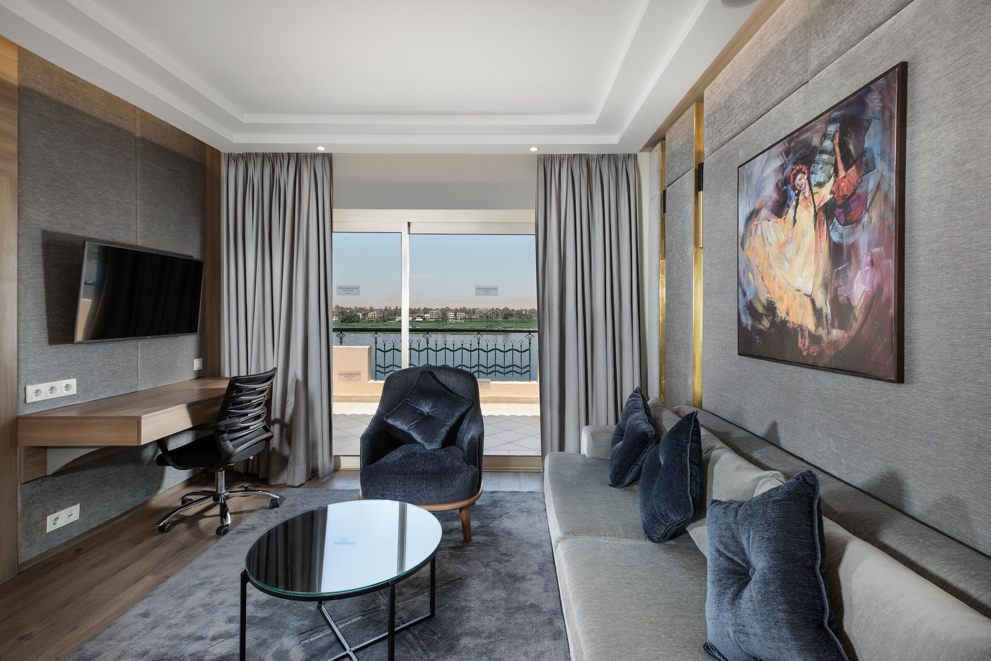 A modern hotel room with a flat-screen TV, a sofa, a chair, a small table, and a large painting.