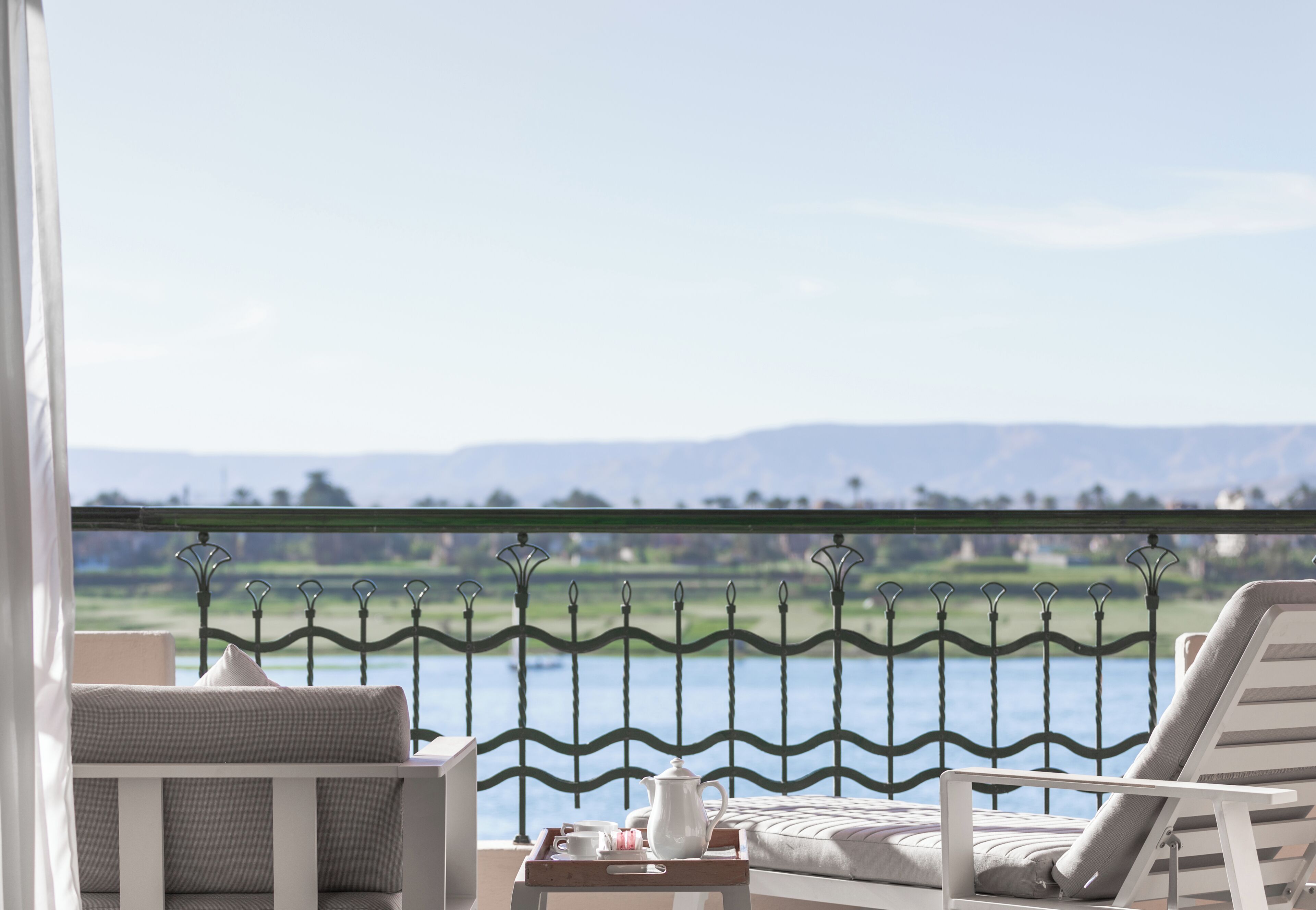 A balcony with a view of a body of water and distant mountains, a white cushioned chair, a small table with a tray of tea items, and a metal railing.