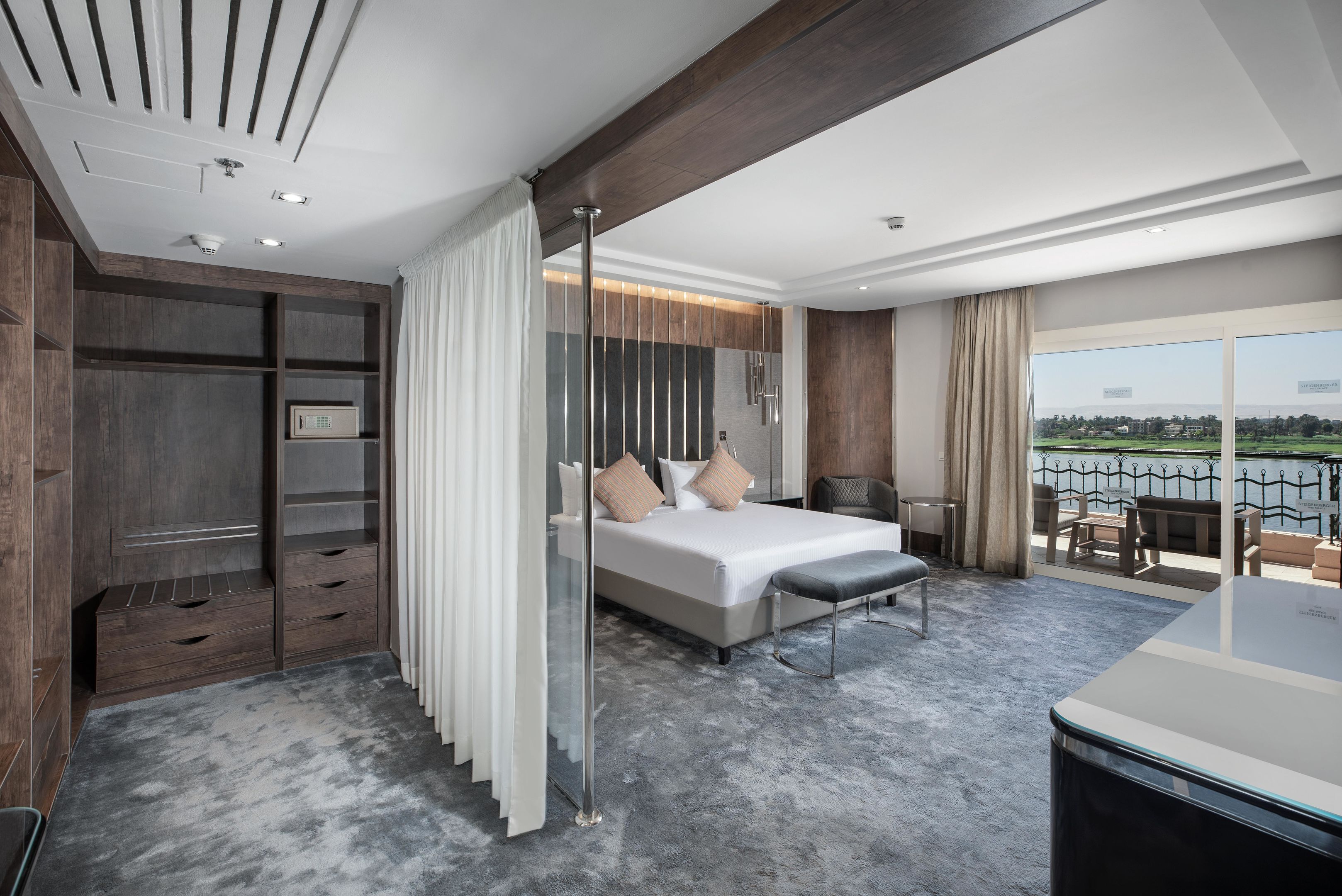 A modern hotel room with a large bed, a glass partition, a desk, and a view of the outdoors.