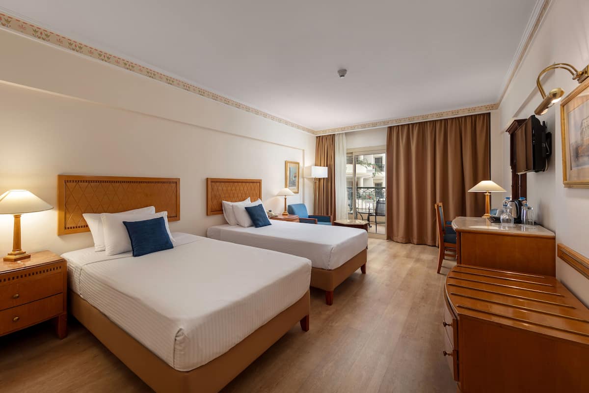 A hotel room with two beds, a desk, a chair, a lamp, and a view of the outside.