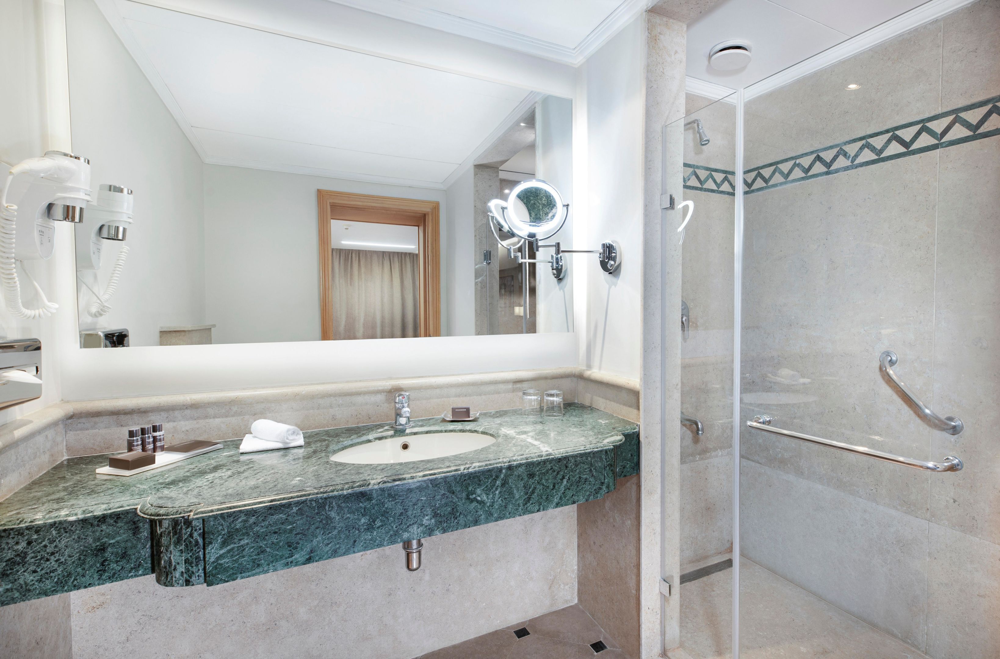 Une salle de bain dotée d’un grand miroir, de deux lavabos, d’une douche avec des barres d’appui et d’un sèche-cheveux fixé au mur.