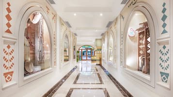 Corredor de hotel com janelas em arco, piso de mármore e vista para um quarto com porta verde.
