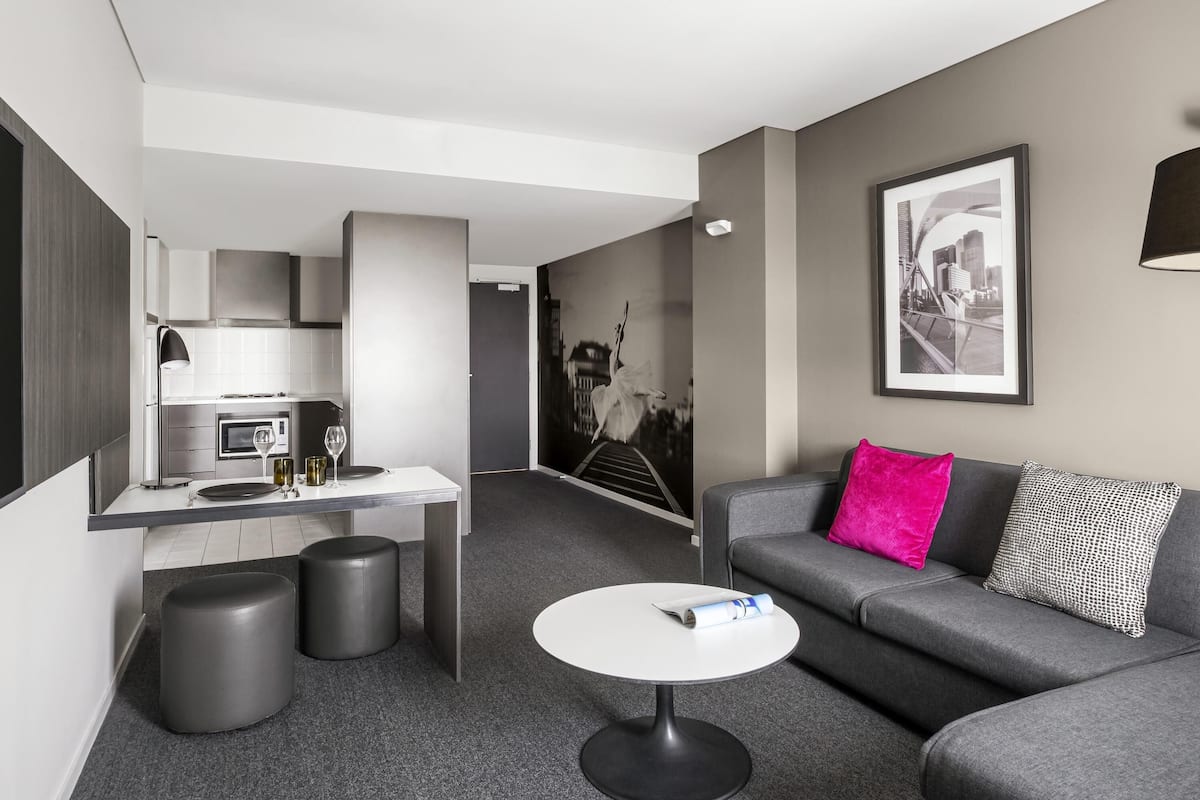 A modern hotel room with a kitchenette, a grey sofa with a bright pink pillow, a round white coffee table, and a cityscape wall art.