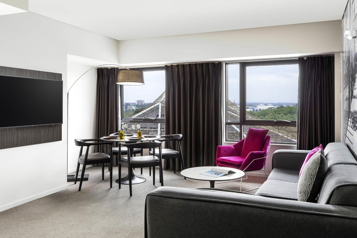 A modern hotel room with a dining area, a sofa, and a large window with a view of a bridge.